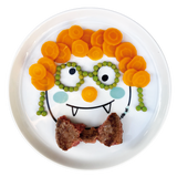 Assiettte pour enfant decoré avec des aliments pour formé le visage rigolo d'un personnage. il y a des carottes qui forment les cheveux, des petits pois pour les lunettes et un steack haché en forme de noed papillon
