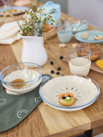 Table en bois ou est posé dessus de la vaisselle contenus un petit déjeuner, en avant plan il y a une assiette en verre avec une base en silicone beige, l'assiette contient une petite tartine en forme de tête
