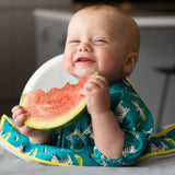 Bébé qui regarde l'objectif et sourit en tenant dans ses mains une tranche de pastèque. Le bébé sembre être assit sur une chaise haute, il a sur lui un bavoir de couleur bleu avec des zèbres.