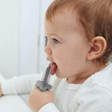 bebe qui porte a sa bouche une brosse à dent de couleur grise, il se brosse la langue. le bebe est de coté sur la photo