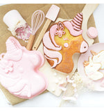 Photo prise en hauteur d'un gâteau en forme de licorne décoré avec un glaçage rose pastel. il y a à coté du gâteau des sablé ainsi que des ustensiles de cuisine rose pastel.