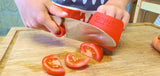 Enfant qui coupe des légumes sur une planche a découper de cuisine. Il utilise le couteau Petit Chef de la marque Opinel avec son couvre doigt rouge de la marque Opinel. Sur la planche a découper il y a des tomates et une carotte. L'enfant découpe une tomate en petite tranche