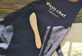 Photo d'un tablier pour enfant bleu avec l'inscription Petit Chef Sohenn, sur le tablier est posé un couteau en bois