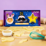 Plan de travail d'une cuisine avec étalé dessus une pâte à biscuit. Sur la pâte est posé une boîte contenant 3 emporte-pièces en forme de tête de lapin, moustache et étoile. Dans leur coffret les découpors sont positionner sur des dessins colorés