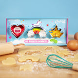 Plan de travail d'une cuisine avec étaler dessus une pâte à biscuit. Sur la pâte est posée une boîte contenant 3 emporte-pièces en forme de cœur, arc en ciel et couronne. Dans leur coffret les découpoirs sont positionnés sur des dessins colorés