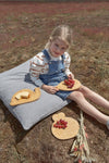 lp Photo d'une jeune fille assise sur un gros coussin dans un champ avec devant elle et sur ses genoux des planches en bois contenant du raisin et de la brioche. La jeune fille est souriante et vêtue d'une salopette en jeans