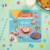 Photo d'un livre de erecette posé sur une table coloré avec des accessoires (petits drapeaus, pic et confettis) dans le theme du livre sont posés dessus. Le livre est intitulé les recettes du monde. La couverture est bleu avec des illustration et photos des recettes.