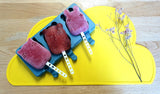 Glace a la framboise dans le moule a glace Frrosties de We might be tiny avec ses botonnets intégrés. Le moule a glace represente 3 formes d'animaux, il est de couleur bleu canrd et est disposé sur un tapis en silicone jaune en forme de nuage. Sur le tapis en silicone il y a également quelques framboises congelées