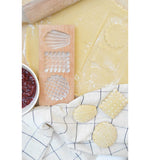 Moule en bois pour réaliser des biscuits fourrés. Le moule est rectangulaire, il est posé sur une table de cuisine, avec une pâte et de la confiture. Il y a 3 empreinte en relief : Gaufrette, petit beurre et madeleine