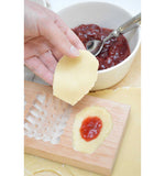 Gros plan sur une main qui pose de la pâte dans un moule a empreinte en bois. Dans le moule il y a déjà de la pâte avec de la confiture à l'intérieur