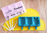 Moule a glace en forme de d'animaux de couleur bleu canard posé sur un tapis en silicone de couleur jaune et en forme de nuage. 5 bâtonnets de glace sont posés à coté du moule. En haut a gauche il y a également le moule a glace de couleur rose et dans son emballage Frosties
