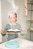 Photo d'une jeune fille dans une cuisine qui porte dans ses mains une grille de cuisson avec dessus un gâteau au chocolat. Sur la table devant est posé un moule en forme de tête d'ours