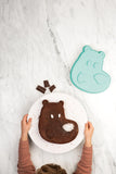 Photo prise par le haut d'une table en marbre avec posé dessus un gâteaux aux chocolat en forme d'ours. Le gâteaux est tenu par les mains d'un enfant qui porte un pull boredaux. Sur le coté de l'assiette est posé le moule qui a servit à la réalisation du gâteau, il est de couleur vert d'eau