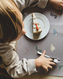 Photo prise en hauteur d"une table avec un enfant assis. L'enfant est devant une part de cheese cake posé dans une assiette et sur un table de couelur gris avec des dessins de dinosaure dessus