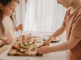 Photo de 2 enfant debout devant une grande planche à découper en bois ou sont posé dessus des morceaux de pommes avec des morceaux de fruits secs.