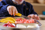 Zoom sur le couteau d'un enfant à manche jaune qui coupe des morceaux de pastèque dans un plateau en inox compartimenté contenant des fruits