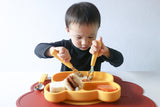 Jeune garçon qui mange à l'aide de couvert aux manche jaune, l'enfant est assis devant une table ou est posé un tapis en silicone de couleur rouille et une assiette a compartiments en forme d'ours de couleur jaune comme les couverts. L'enfant semble concentré sur son assiette