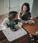 Photo de 2 enfant entrain de manger. ils sont assis devant une table à manger en bois foncé avec des set de table posés devant eux. Ils portent tous les 2 le même bavoir de couleur olive. les tons de la photo sont épurés et de couleur scandinave
