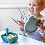 Photo d'un jeune garçon au yeux bleu, souriant. il est assit à table devant un bol de couleur bleu contenant des pâtes.