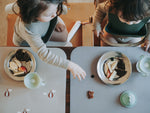 Photo prise en hauteur de 2 enfant entrain de manger, il y a de posé devant eux 2 set de table de couleurs differentes avec des assiettes en inox contenant de la nourriture