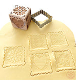 Pâte à sablé étalé avec des biscuits en forme de noel découpé dessus. Au dessus de la pâte il y a l'emporte pièce en inox qui a permeis de découper les biscuits ainsi que son tampon en bois en forme de dé
