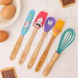 Ensemble de 5 ustensiles de cuisine pour enfant. Ils sont posé sur une table de cuisine blanche, avec autour des gâteaux et des oeufs.. Les manche sont en bois gravé avec la marque Chefclub et les têtes en silicone coloré avec des illustrations de personnages. il y a un fouet, une spatule, une cuillère, un pinceau et une maryse