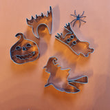 Emportes pieces halloween. Il y en a 4, un en forme de citrouille, chat qui a peur, fantôme et sorcière. Les emporte piece sont en aluminium et posés sur un fond orange. Sur le coté droit de la photo est posé une araignée