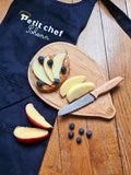 Photo d'un tablier bleu avec posé dessus une planche à repas en bois en forme de herisson. Sur la planche il y a un couteau avec une lame en acier dentellée et un manche en bois. Sur la planche il ya également une tartine avec des fruits dessus et des morceaux de pommes à coté. Sur le tablier il y a une inscription : Petit chef Sohenn