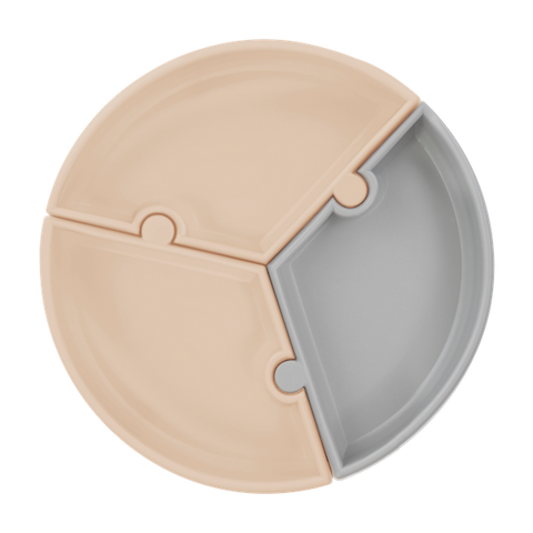 Assiette en silicone en forme de puzzle. Elle est composée de 3 compartiments qui s'emboitent comme des pièces de puzzle. Il y a 2 compartiment de couleur beige et un gris.