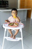 Jeune enfant entrain de manger dans une cuisine, il est assit sur une chaise haute équipé d’un bavoir tablier qui s’accroche directement sur la chaise haute. Il est de la marque Tidy Tot