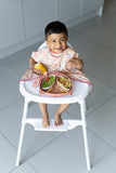 Jeune enfant entrain de manger dans une cuisine, il est assit sur une chaise haute équipé d’un bavoir tablier qui s’accroche directement sur la chaise haute. Il est de la marque Tidy Tot