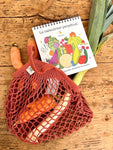 Filet de course de couleur rouge, contenant des carottes et un poireau. il est posé sur un plan de travail en bois. un calendrier perpétuel illustré de fruit et légumes rigolos sort également du sac à provisions