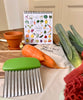 Calendrier perpétuel posé sur une table en bois, il est ouvert sur le mois d'octobre. des fruits et légumes rigolos sont représenté. Sur la table sont disposés une carotte et des poireaux avec un coupe légumes et un petit pot en terre cuite