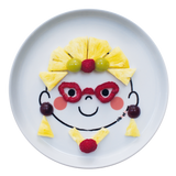 Assiette en porcelaine blanche avec une illustration de tête de bonhomme rigolos au centre. Elle est de la marque Cibolo , indiqué sur l’oval du visage. L’assiette permet de créer des repas rigolos en animant le personnage. Elle est composé de morceaux de fruits, representant une femme avec des lunettes et des bijoux