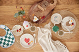 Photo prise en hauteur d'une table à manger avec 2 sets de tables en osier ou sont posés de la vaisselle pour enfant colorés avec des illustration de coeur souriant et d'emoji. La vaisselle contient des tartine et des fruits