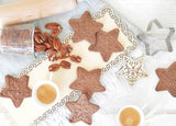 Biscuit en forme d'étoile avec des motifs dessus, ils sont disposés sur une table avec une boît en verre contenant des noix de cajoux. Il y a également l'emporte-pièce et le tampon en bois qui ont permis de réaliser les sablés