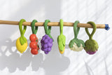Ensemble de plusieurs hochet suspendu à un morceau de bois. Les hochet ont un anneau et leur embout représentent des fruits et légumes, il y a un citron, des tomates cerises, du raison, un épi de mais, un chou fleur et un artichaut