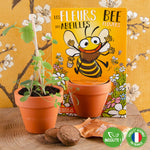 kit de jardinnage pour enfant pour faire pousser des fleurs d'abeilles. Le kit est présenté dans un emballage de couleur jaune avec des illustration de fleurs et d'une abeille rigolotte. Le kit est posé sur une table en bois devant un papier peint de couleur jaune avec des fleurs de cerisier