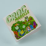 Photo de la couverture  du livre pour enfant Croc Marelle de Manon Brzostek. La couverture est illustrée par des dessins d'une petote fille allongé dans de l'herbe avec des fruits et légumes rigolos tout autour.