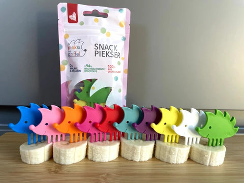Ensemble de 10 pics alimentaires de différentes couleurs avec des formes d'hérissons. Ils sont posés sur des morceaux de banane, les uns derrière les autres. Derrière, il y a le paquet d'emballage des pics alimentaires de la marque Pieksi und Griffel.