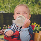 Photo d'un bébé blond, qui tient dans ses mains et dans sa bouche une tasse avec paille. il est assit sur une chaise haute et est penché légèrement sur le coté, en regardant la personne qui le prend en photo