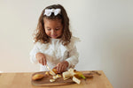 Jeune fille qui coupe des morceaux de madeleine, elle est devant une table avec une planche en bois. Elle utilise un couteau pour enfant de la marque kiddikutter