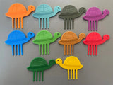 Ensemble de 10 pics alimentaires de différentes couleurs avec des formes de tortues. ils sont posés sur un fond gris à plat les uns devant les autres