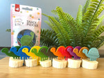 Ensemble de 10 pics alimentaires de différentes couleurs avec des formes de tortues. Ils sont posés sur des morceaux de banane, les uns derrière les autres. Derrière, il y a le paquet d'emballage des pics alimentaires de la marque Pieksi und Griffel.
