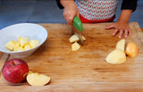 Petites mains d'enfants entrain de découper des morceaux de pommes à l'aide d'un decoupe legume en acier avec une poignée verte. La pomme est découpée sur une planche a découper en bois avec dessus des pommes et un saladier blanc avec des morceaux déjà coupés 