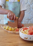 main d'enfant qui coupe des morceaux de pastèque à l'aide d'un coupe legumes en bois gravé de la marque Skagfa. L'enfant porte un talier bleu clair avec des illustrations de zèbre dessus. Devant la planche à découper est posé un petit bol avec des morceaux de fruits à l'intérieur