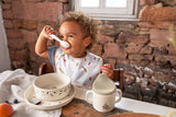 Photo d'un jeune garçon assis devant une table avec de la vaisselle assortie devant lui. il est entrain de manger. Derrière lui il y a un mur en brique rouge