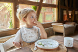 Jeune fille assise à table, qui est entrain de manger. Elle a un tablier assorti avec la vaisselle qui est posé sur la table devant emme. La photo semble prise dans un chalet en bois