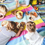 Photo prise en hauteur d'un pique nique d'enfants. les enfants sont assis sur un plaid coloré avec plusieurs assiettes et planches en bois posé dessus avec différents aliments.