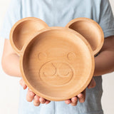 Assiette en bois pour enfant en forme de tête d'ours, elle possède 3 compartiments. L'assiette est tenu dans les main d'un enfant